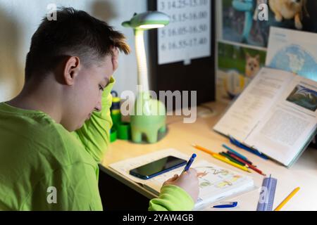 Un garçon fait ses devoirs à son bureau. Avec une main sur le front, il écrit dans son cahier avec l'autre. Il y a un smartphone à proximité Banque D'Images