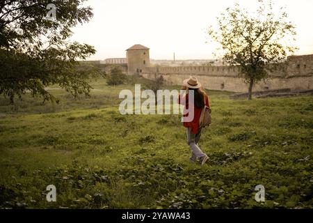 Vue arrière de la femme touriste avec sac à dos marchant dans la nature sur fond de vieille forteresse au coucher du soleil dans la soirée. Voyage, tourisme et tourisme co Banque D'Images