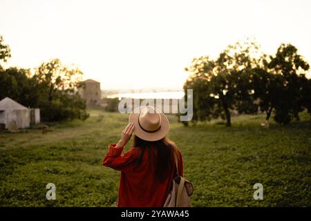 Femme en chapeau marchant vers la forteresse historique au coucher du soleil, entourée de verdure et d'arbres. Scène paisible et sereine capture la nature et l'histoire merg Banque D'Images