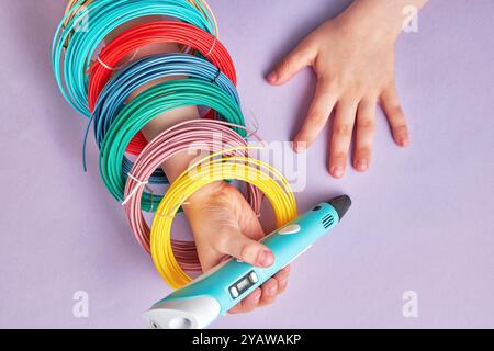 Childs Hand tient un stylo 3D avec des filaments colorés à proximité. Les couleurs vibrantes sont prêtes à être utilisées pour créer des designs uniques et imaginatifs Banque D'Images