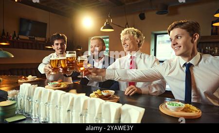 Groupe d'hommes d'affaires en costumes élèvent leurs verres de bière, griller à la journée réussie. L'éclairage chaleureux du bar ajoute une ambiance festive. Banque D'Images