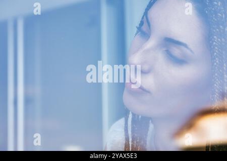 Jeune femme avec des tresses se tient près d'une fenêtre perdue dans la contemplation profonde, exsudant la sérénité et le calme alors qu'elle rêve dans la solitude paisible, illumine Banque D'Images