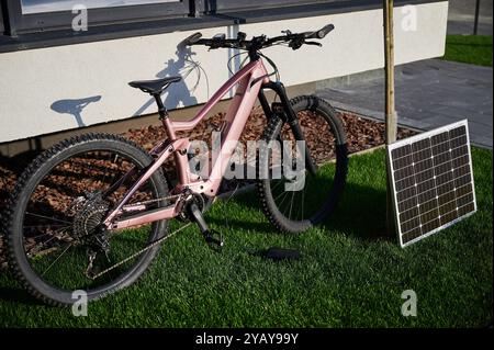 Vélo de montagne électrique rose garé sur une pelouse herbeuse, connecté à un panneau solaire pour la recharge. Vélo dispose de pneus robustes et positionné à côté du bâtiment, avec son ombre projetée sur le mur. Banque D'Images