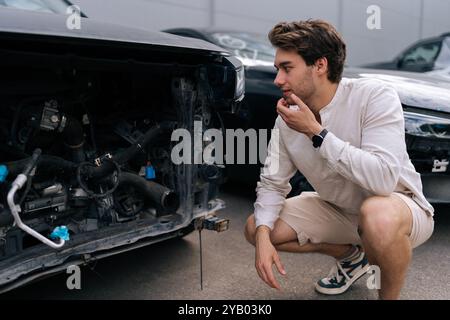 Stressé propriétaire de voiture masculin squatting et inspectant les dommages à la voiture après l'accident. Conducteur frustré regardant la voiture endommagée. Banque D'Images