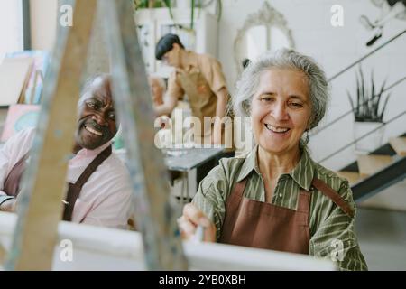 Femme aux cheveux argentés souriant largement tout en appréciant le processus de dessin, son mari afro-américain regardant sa peinture Banque D'Images