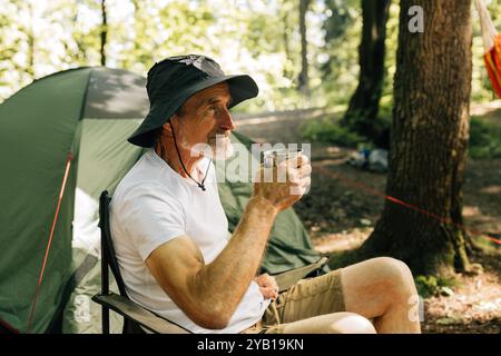 Vue de côté d'un homme âgé avec la tasse assise dans une chaise contre la tente dans la forêt Banque D'Images