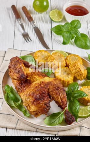 la moitié de poulet rôti avec des pièces d'épis de maïs et des feuilles de basilic frais sur une assiette sur une table en bois blanc avec couverts, citron vert et sauce chili doux, verticale Banque D'Images