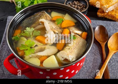 Soupe claire de brochet avec légumes, persil et épices dans un pot rouge sur une table en bois noir avec des cuillères en bois et du pain grillé, gros plan Banque D'Images