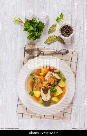 Soupe claire de brochet avec légumes, persil et épices dans un bol blanc sur une table en bois blanc avec cuillère, feuilles de laurier et grains de poivre, vue verticale d'abo Banque D'Images