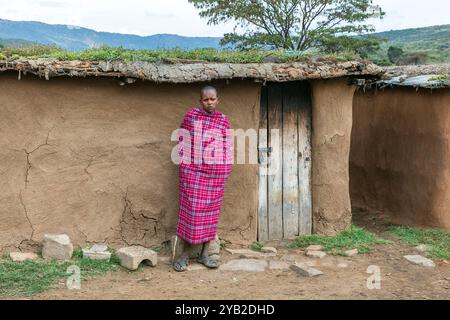 Jeune garçon Masai debout devant une maison traditionnelle Masai faite de bouse de vache, boue et branches de bois, Masai Mara, Kenya, Afrique Banque D'Images