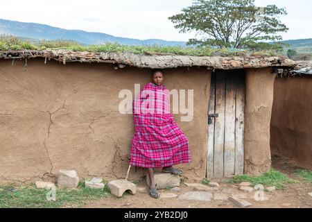 Jeune garçon Masai debout devant une maison traditionnelle Masai faite de bouse de vache, boue et branches de bois, Masai Mara, Kenya, Afrique Banque D'Images