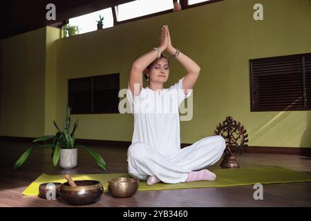 Femme pratiquant la méditation dans le yoga Kundalini avec un geste namaste accueillant et les mains levées, assise dans une position confortable de lotus sur tapis dans la chambre Banque D'Images