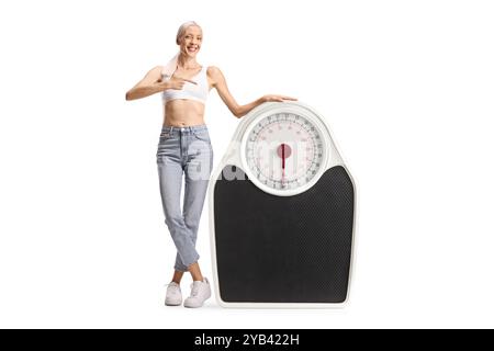Femme heureuse en jeans et crop top debout pointant à une grande échelle de poids isolé sur fond blanc Banque D'Images
