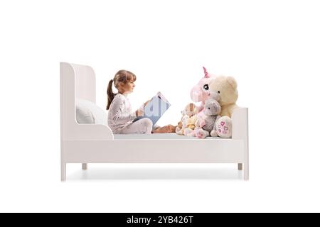 Enfant assis dans un lit avec des jouets pour animaux et lisant un livre isolé sur fond blanc Banque D'Images