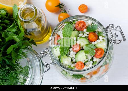 Salade de couscous frais dans un pot en verre avec concombres, tomates, herbes, citron avec des ingrédients sur fond blanc. Salade de taboulé. Végétarien en bonne santé Banque D'Images