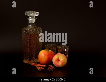 Liqueur pêche-pomme à la cannelle et anis sur fond noir, alcool fort maison dans un bol en cristal, fruits mûrs et épices à proximité. Gros plan. Banque D'Images