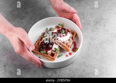 Serveur servant une assiette blanche avec des gaufres belges sucrées avec de la crème et de la confiture de cerises sur une table grise dans un café Banque D'Images