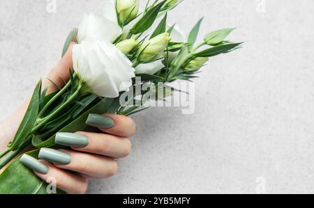 Une main avec de longs ongles élégants tient un bouquet de fleurs blanches fraîches. Les verts et les blancs doux contrastent magnifiquement avec un dos texturé léger Banque D'Images