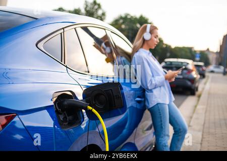 Gros plan sur la voiture électrique se chargeant au bord de la route, tandis qu'une femme se tient à proximité portant des écouteurs, écoutant de la musique Banque D'Images