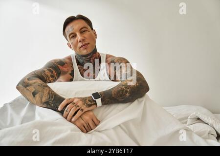 Un jeune homme avec des tatouages frappants est assis confortablement dans son lit, exsudant confiance et style détendu. Banque D'Images