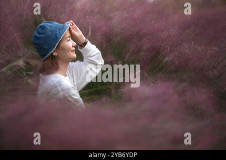 Sourire jeune femme asiatique tenir le chapeau bord, vue de côté. entouré d'herbe muhly de hairawn pourpre (muhlenbergia capillaris) Banque D'Images