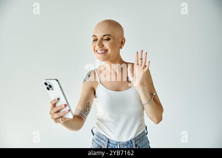 Une jeune femme chauve sourit brillamment et fait des vagues en se connectant avec des amis en ligne depuis sa maison. Banque D'Images