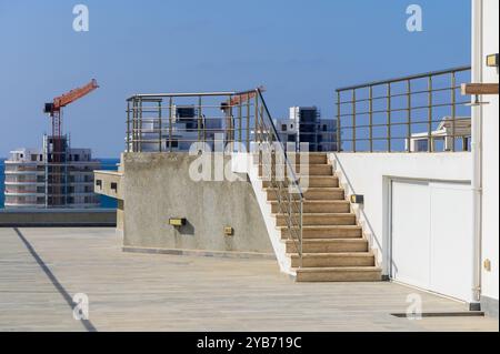Ce toit présente des escaliers élégants menant au haut, entourés de bâtiments contemporains et de grues de construction sous un ciel bleu clair, évoquant t urbain Banque D'Images