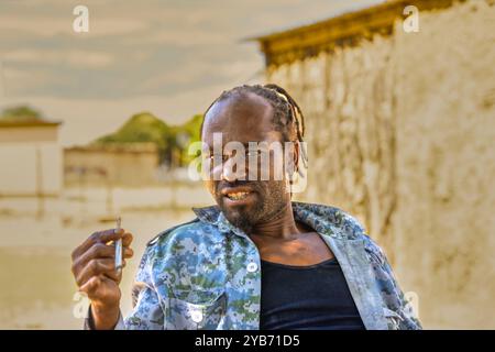 homme africain du village avec des dreadlocks debout devant la maison fumant de la marijuana Banque D'Images