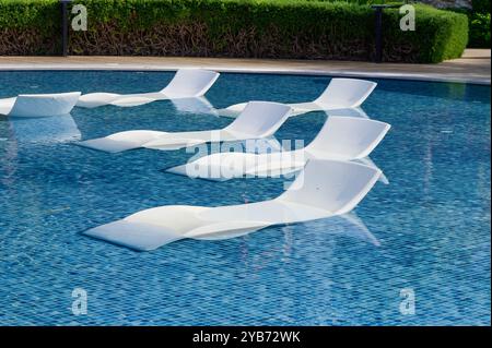 Des chaises longues sont partiellement immergées dans une piscine d'un bleu clair, invitant à la détente tandis que la végétation luxuriante encadre le cadre paisible sous la lumière du soleil Banque D'Images