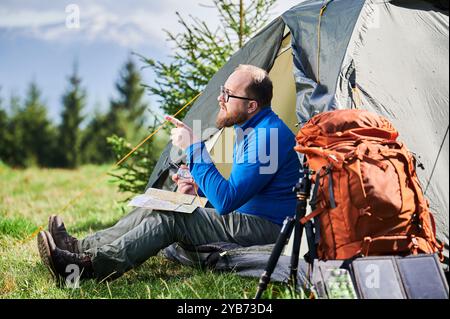 L'homme est assis dans l'entrée de la tente touristique, pointe dans la distance tout en étudiant la carte. À côté de lui sac à dos orange, équipé d'un trépied et d'un panneau solaire, placé sur fond pittoresque d'arbres et de montagnes. Banque D'Images