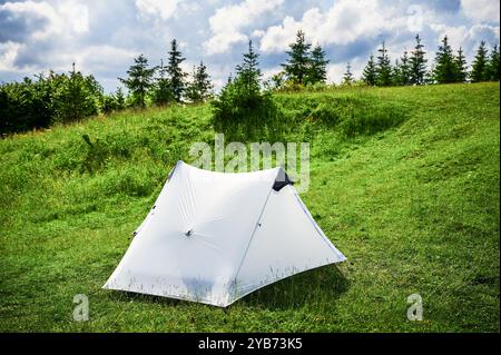 Tente touristique blanche au soleil, au sommet d'une colline herbeuse, entourée de jeunes pins. Vue panoramique sur les montagnes luxuriantes et vallonnées sous un ciel bleu vif avec des nuages dispersés, créant un endroit de camping idyllique. Banque D'Images