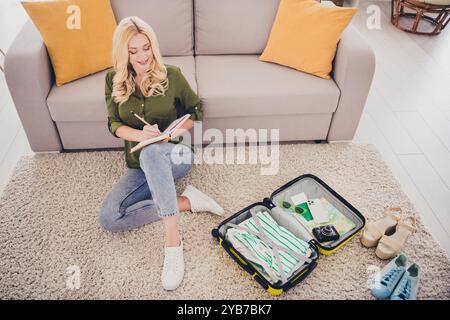 Photo d'adorable jolie femme assise sur le sol écrivant des plans pour un futur voyage de week-end organisant valise à l'intérieur Banque D'Images