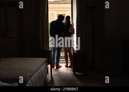 Silouete d'Un couple face à face avec une valise rouge dans une chambre d'hôtel avec la fenêtre ouverte. À l'extérieur, vous pouvez voir un architecte européen classique Banque D'Images