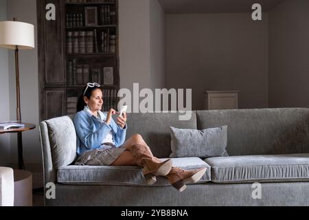 Une belle femme adulte aux cheveux noirs regarde son smartphone calmement et détendue. Elle est assise confortablement sur un canapé gris dans une chambre avec d classique Banque D'Images