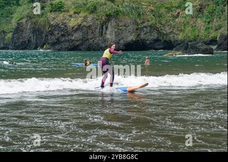 Une jeune surfeuse perfectionne son équilibre sur une petite vague lors d’une leçon de surf à la plage de Seixal, avec des falaises luxuriantes en toile de fond Banque D'Images