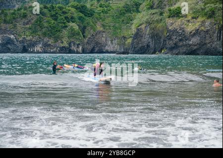 Une jeune surfeuse s'entraîne debout sur sa planche pendant une leçon d'école de surf à la plage de Seixal, avec des camarades et des falaises luxuriantes en arrière-plan Banque D'Images