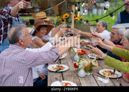 Groupe d'amis seniors levant des verres dans un toast lors d'un dîner en plein air. Les personnes âgées appréciant le repas et le vin dans le jardin avec des fleurs et St Banque D'Images