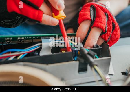 Électricien en gants travaille méticuleusement sur une carte de circuit imprimé, connectant les fils avec un tournevis. L'image en gros plan montre des mains habiles fixant un appareil cassé avec précision Banque D'Images