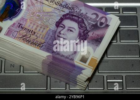Une pile de notes de vingt livres repose sur un clavier d'ordinateur portable argenté, avec la note du haut montrant l'image de la reine Elizabeth II Banque D'Images