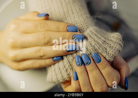 Dans un cadre intérieur confortable, les mains avec vernis à ongles bleu arborant des clous argentés tiennent doucement un pull tricoté. La scène capture un moment de rela Banque D'Images