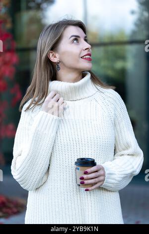 belle fille dans un pull tricoté blanc avec une tasse de café dans la rue sur fond de feuilles lumineuses d'automne, la vie urbaine en automne Banque D'Images