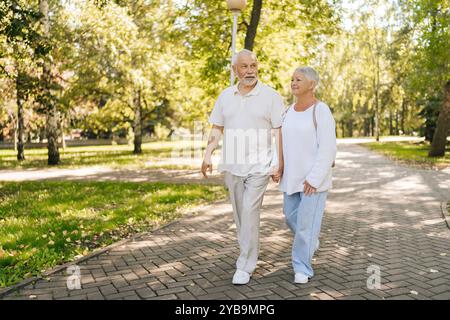 Portrait en pied d'un couple senior profitant d'une promenade tranquille dans un parc verdoyant, se tenant les mains entrelacées, partageant un moment tendre le jour ensoleillé Banque D'Images