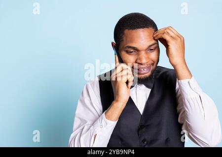 Chasseur afro-américain effectuant des réservations de chambre sur téléphone intelligent, partageant des informations sur les équipements de l'hôtel sur fond bleu. Le jeune portier répond aux questions sur la caméra. Banque D'Images