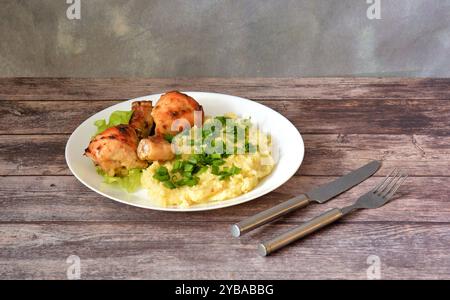 Cuisses de poulet cuites au four avec purée de pommes de terre et oignons verts hachés sur une table en bois clair, couverts à proximité. Gros plan. Banque D'Images