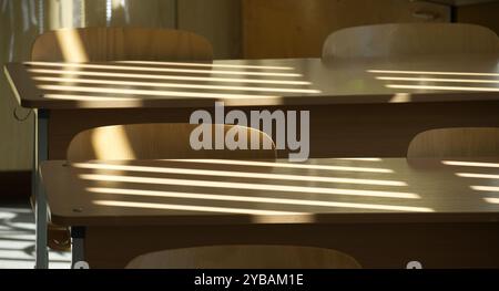 Salle de classe vide avec bureaux et chaises en bois avec lumière du soleil projetant des lignes d'ombre à travers les stores, créant une atmosphère sereine et calme. Banque D'Images