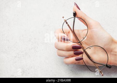 Une main avec de longs ongles Bordeaux tient gracieusement une paire de verres ronds au-dessus d'une surface texturée légère, éclairée par une lumière naturelle douce. Banque D'Images