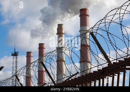 Les cheminées d'usine fument de la fumée épaisse et de la vapeur derrière une clôture avec du fil de fer barbelé Banque D'Images