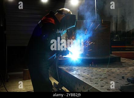 Métallurgiste dans le masque de sécurité et les gants utilisant la torche de soudage pour joindre les côtés de la construction métallique dans l'atelier à l'usine. Thème industriel Banque D'Images