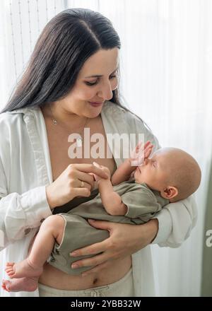 Maman se tient près de la fenêtre et tient le bébé nouveau-né dans les bras. Maman touche doucement la main du fils endormi. Soins et soutien, sommeil et protection. Amour et relatio Banque D'Images