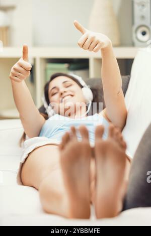 Jeune femme est confortablement allongée sur son canapé à la maison, écoutant de la musique avec des écouteurs, donnant un geste pouce levé, paraissant détendue et heureuse Banque D'Images